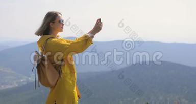 穿着黄色雨衣的女孩在智能手机上拍照，站在山顶、蓝天和群山上
